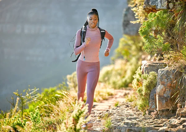 Young fit and active african american woman hiking and jogging through the mountains on a sunny summer day. Black woman running alone exercising and carrying a backpack while exploring nature looking