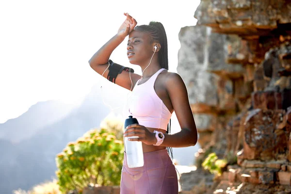 Young african american woman wearing earphones listening to music while taking a water break on a mountain in the morning. Confident fit black female resting after a run alone in nature.