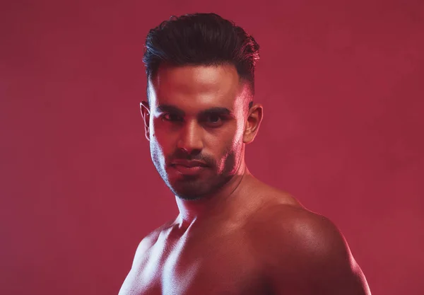 One handsome young indian man posing topless against a red studio background. Serious masculine guy looking confident and charming with muscular bare body.