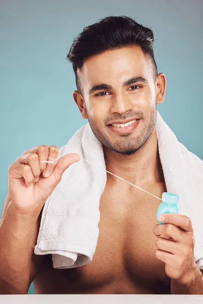 Portrait of one smiling young indian man with towel around neck flossing his teeth after a shower against a blue studio background. Happy guy cleaning his mouth for better oral and dental hygiene