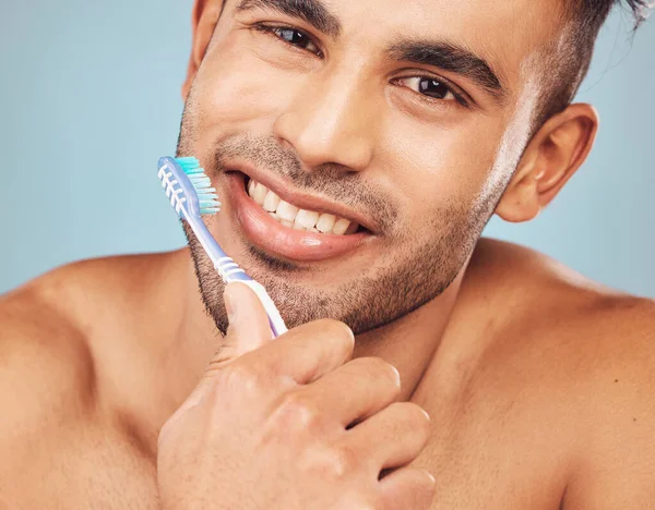 Portrait of one smiling young indian man brushing his teeth against a blue studio background. Handsome guy grooming and cleaning his mouth for better oral and dental hygiene. Brush twice daily to