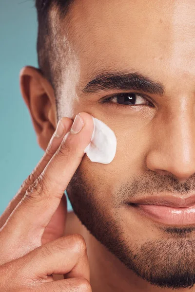 Closeup of one young indian man applying moisturiser lotion to his face while grooming against a blue studio background. Handsome guy using sunscreen with spf for uv protection. Rubbing facial cream