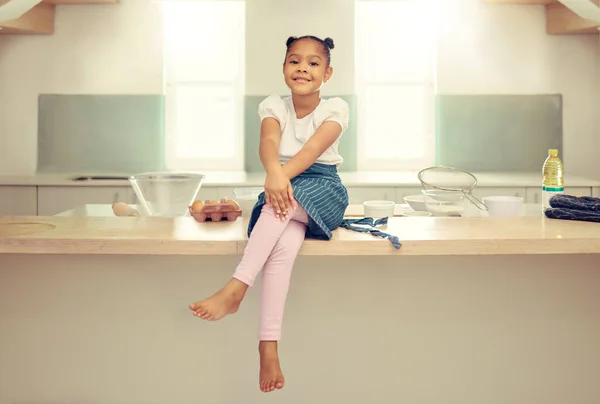 Portrait of a cute young mixed race girl sitting on the kitchen counter with an apron and smiling. Little hispanic girl smiling and sitting alone while baking home. She enjoys cooking in the kitchen.