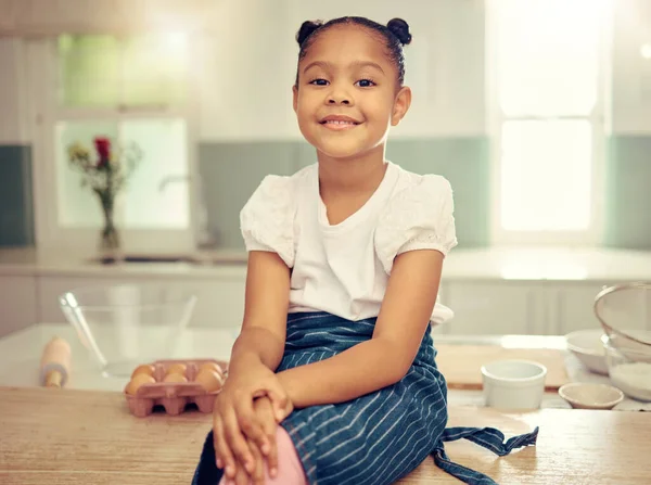 Portrait of a cute young mixed race girl sitting on the kitchen counter smiling and wearing an apron looking thoughtful. Little innocent hispanic girl smiling and sitting alone after baking in the