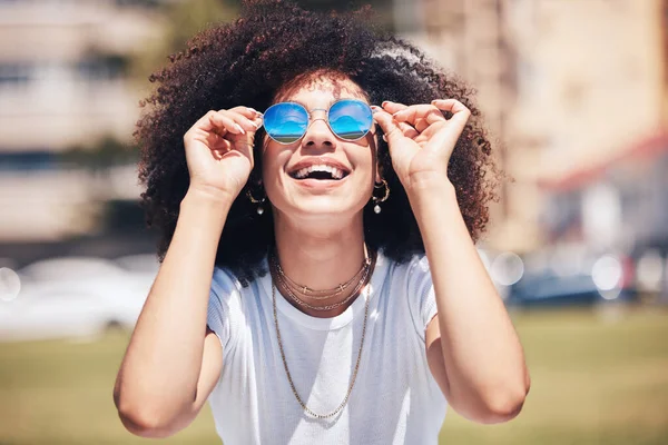 Cool, happy hispanic woman wearing sunglasses outside. Cheerful young woman with a curly afro wearing trendy, stylish sunglasses while enjoying a summer day at the park outside. Young woman laughing.