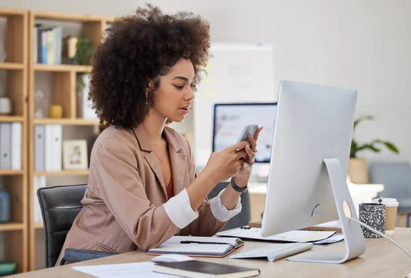 One young busy African American woman using a desktop and cellphone at a desk in her office job. Black woman with an afro checking social media at her job. Mixed race woman focused on texting.