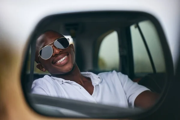 One African American man looking in the car mirror while taking a roadtrip. Smiling black man enjoying the weekend and taking a trip in a vehicle while wearing sunglasses.