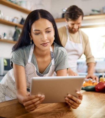 Erkek arkadaşı arka planda yemek pişirirken genç ve güzel bir kadının dijital tablete göz atması. Evde yemek pişirirken internetten yemek tarifi arayan genç İspanyol kadın.