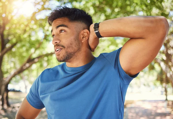 Fit athletic man smiling while stretching his arms during a workout at the park. Hispanic man doing warm-up exercises outdoors on a sunny day. Athletic man taking a break from a fitness routine.