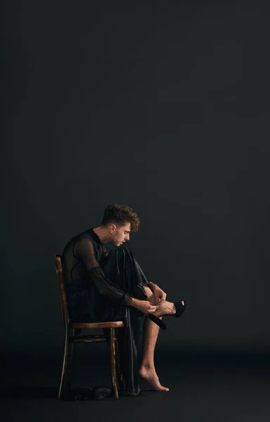 Full body of one handsome caucasian gay man isolated against a black background in a studio and sitting while wearing high heels. Transvestite wearing a dress. Freedom, expression in LGBTQ community.