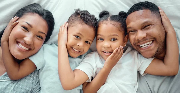 Portrait of happy mixed race family with two children relaxing and lying together on a bed at home, from above. Little brother and sister with their hands on each others faces while bonding with their