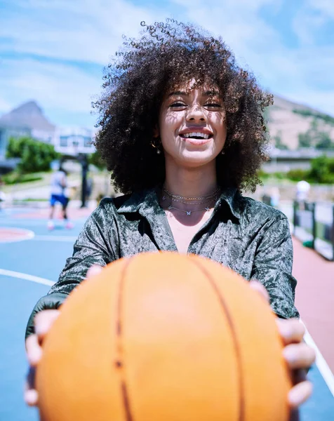 Happy young hispanic woman about to workout holding a basketball while standing on a court. Portrait of smiling girl about to play basketball holding and showing the ball outside