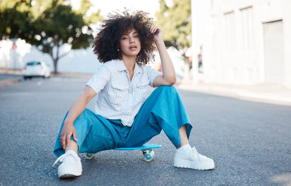A young female mixed race woman skate sitting on a skateboard looking cool and confident with great style in a street outside. Hispanic hipster woman with curly afro hair style in a cool outfit