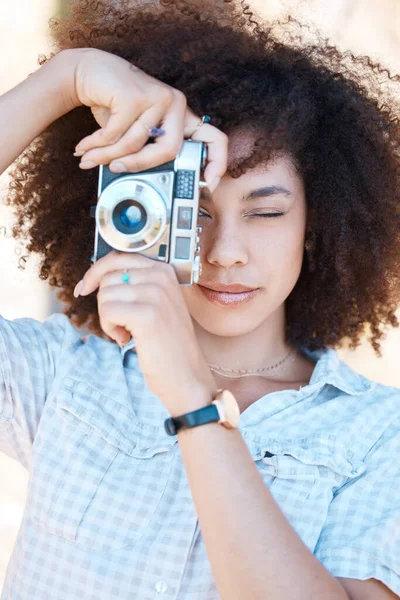 Young mixed race woman with curly hair taking creative photos on a vintage retro film camera. One female photographer looking in viewfinder while capturing pictures as a hobby or profession on a shoot