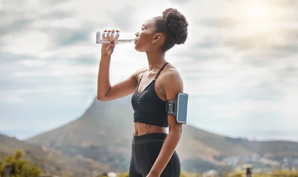 One young african american woman taking a break to drink some water while exercising outdoors. A beautiful and fit mixed race female athlete drinking from a bottle while working out in nature.