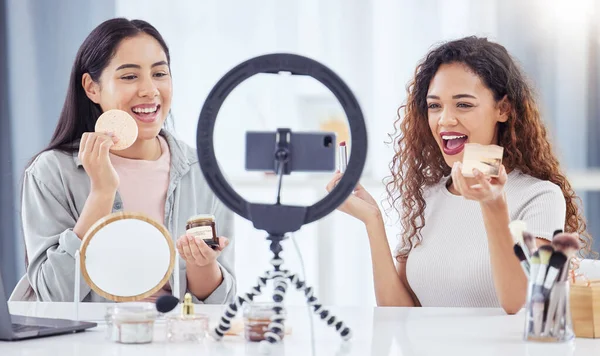 Two mixed race women recording a makeup tutorial for their beauty blog with a phone while sitting at home. Diverse young influencers live streaming their cosmetic review and recommendation online.