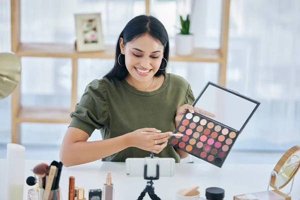One beautiful mixed race creative woman sitting alone in a studio and using her cellphone to vlog while showing her eyeshadow palette. Happy hispanic makeup artist filming a tutorial for the internet.