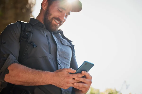 Happy caucasian man using smartphone while carrying a backpack and hiking alone in the mountains on a sunny day. Smiling fit and active man using mobile app or messaging while in a nature environment.