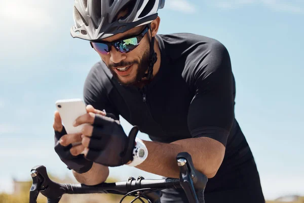 Happy young cyclist wearing helmet and glasses while using his smartphone and sitting on his bike. Fit sportsman using app or sending a message on mobile phone while cycling outdoors.