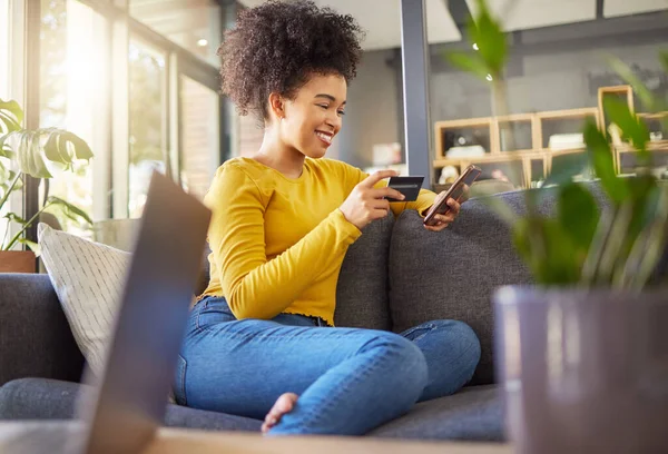 Young happy mixed race woman using a credit card and phone alone at home. Cheerful hispanic female with a curly afro making an online purchase with a debit card and cellphone while sitting on the