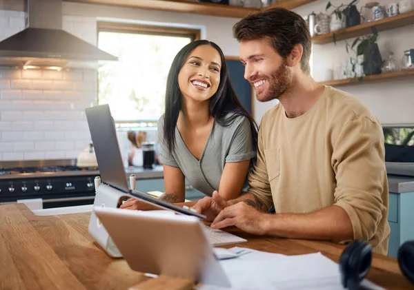 Happy young caucasian man working on laptop in the kitchen at home while his wife stands next to him and smiles while he looks at screen. Happy young interracial couple surfing the internet and