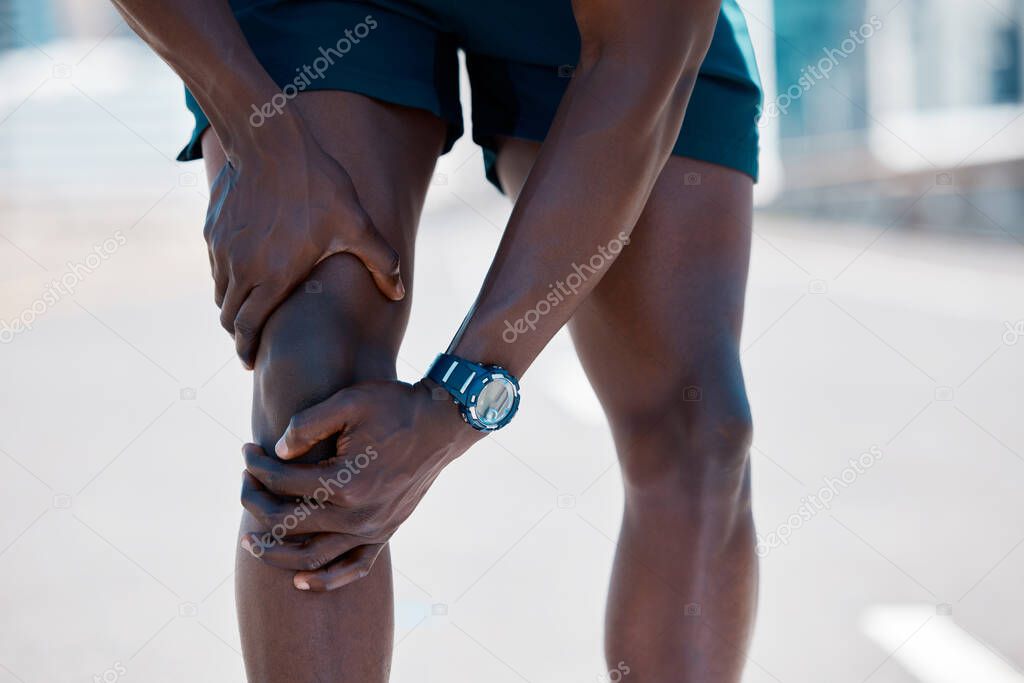 African american athlete taking a break from running to touch his knee and inspect an injury. Fit, sporty, healthy man feeling pain in his knee joint during a jog in the city. Athlete working out.