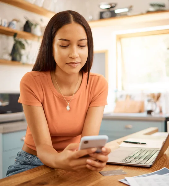 Young beautiful happy mixed race businesswoman holding and using a phone while working from home. One content hispanic woman using social media on her cellphone while using a laptop.