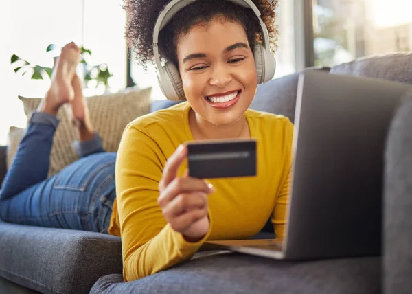 Young happy mixed race woman using a credit card and laptop while wearing headphones and listening to music alone at home. Cheerful hispanic female with a curly afro making an online purchase with a