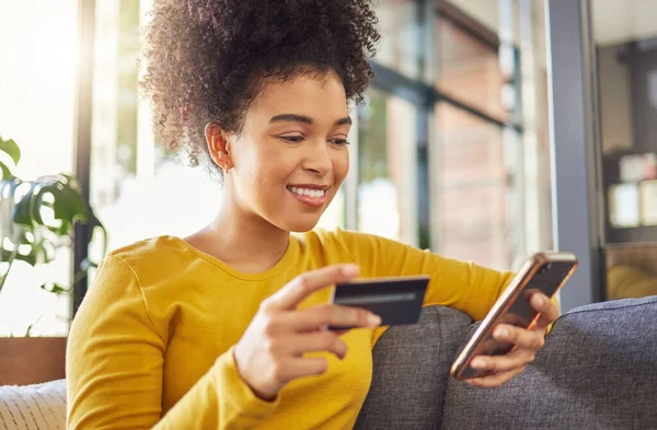 Young happy mixed race woman using a credit card and phone alone at home. Content hispanic female with a curly afro making an online purchase with a debit card and cellphone while sitting on the couch