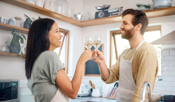 Young interracial couple sharing toast with wine glasses while wearing aprons and cooking together in kitchen at home. Young couple enjoying romantic homemade dinner for date night at home.