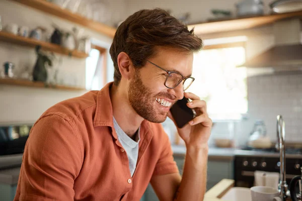 Young content caucasian businessman wearing glasses on a call using a phone while working at home alone. One happy male businessperson smiling and talking on a cellphone while working in the kitchen