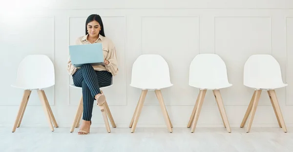 Full body indian businesswoman sitting alone and using a laptop to browse the internet while waiting for an interview. Creative candidate searching online for vacancies. Staying connected and online.