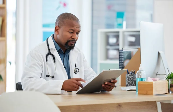 Mature african american doctor checking a patients chart. Focused gp reading a chart on a clipboard. Serious doctor reading a patients report. Professional physician reading a medical document.