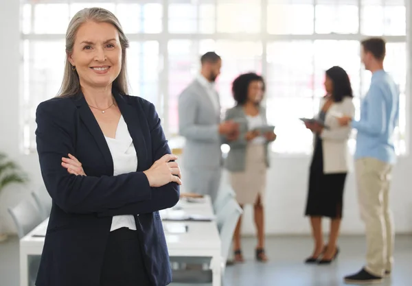 Mature caucasian businesswoman standing with her arms crossed while in an office with colleagues. Happy female boss In a meeting with coworkers.
