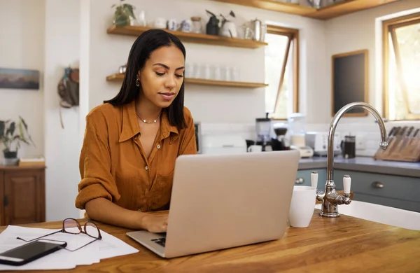 Young content mixed race businesswoman going through paper and bills while typing on a laptop at home. Serious hispanic female businessperson typing an email on a laptop while working from home.