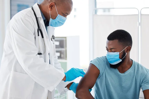 Doctor injecting a patient with the covid vaccine. African american doctor holding a needle filled with the cure. Medical gp giving a patient the corona cure. Doctor and patient wearing masks.