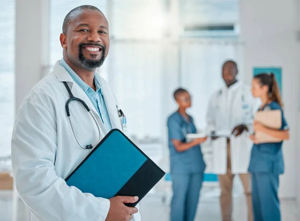 Portrait of a mature african american male doctor holding a folder working at a hospital with colleagues. Expert medical professional smiling ready for work at a clinic with coworkers.