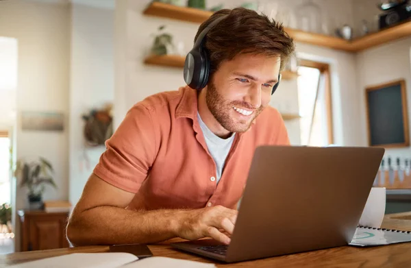 Young cheerful caucasian businessman wearing headphones and listening to music while using a laptop at home alone. One content male businessperson typing on a laptop while working in the kitchen at