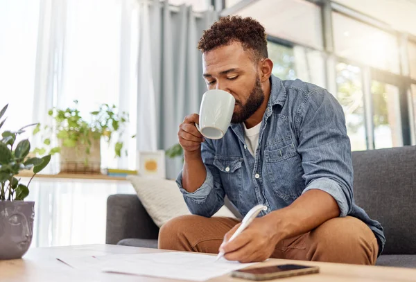 Young mixed race man working on filling out forms while drinking coffee at home. One hispanic person drinking a cup of tea while planning alone in the lounge at home.
