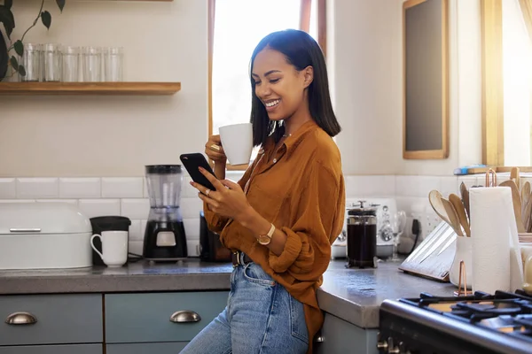 Young happy mixed race woman drinking a cup of coffee while using her phone alone in the morning in the kitchen. One hispanic female smiling and enjoying a cup of tea while using social media on her