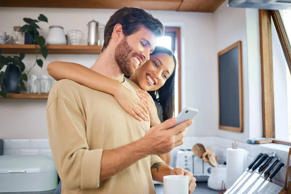 Happy young interracial couple being loving and affectionate at home. Young man using his smartphone and holds coffee cup while his girlfriend embraces him from behind. Browsing social media or