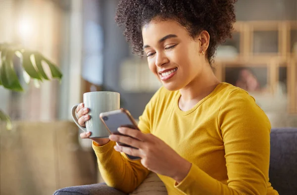 Happy mixed race woman drinking a cup of coffee and typing a message on a phone at home. One content hispanic female with a curly afro using social media on a cellphone while relaxing on the couch at