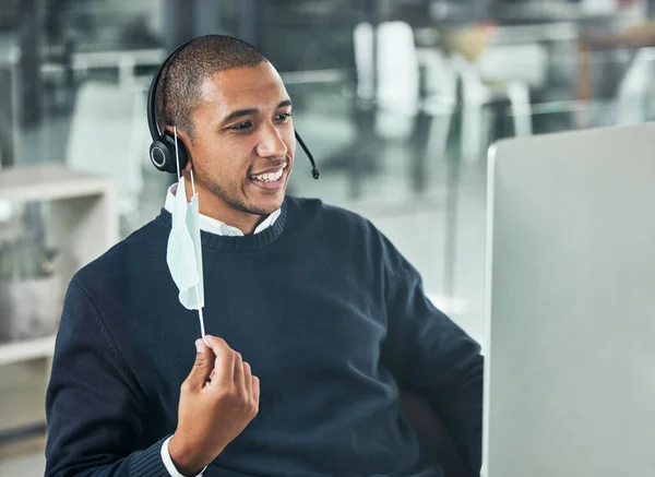 Young male call center agent smiling and wearing a headset and mask working on a computer in an office at work. Customer service, support and sales. Giving advice and helping. Answering calls.