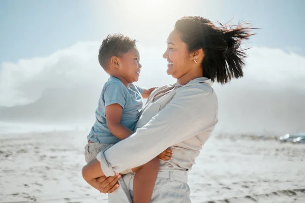 Loving young mixed race mother holding her adorable little son as they looking into each others eyes on a windy day at the beach. Mother and son spending time and having fun together while on holiday.