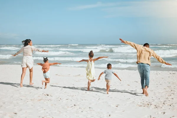 Rear view of a playful mixed race family with three children running on the beach with arms outstretched. A big family spending time and enjoying holiday by the sea.