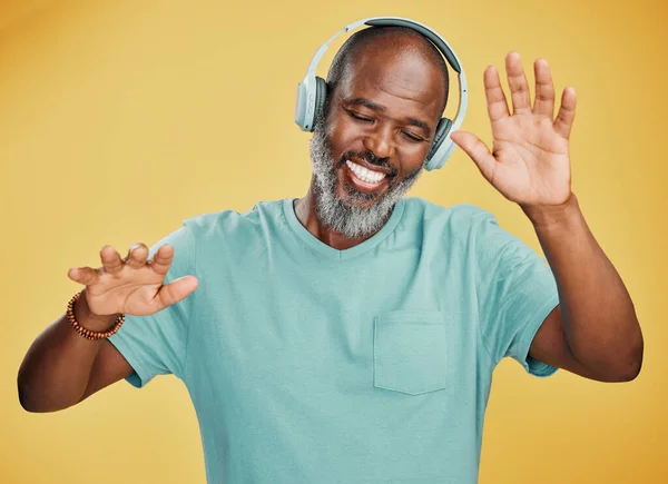 Happy mature african man dancing while listening to music with headphones against a yellow studio background. Carefree senior black man with grey beard celebrating to and enjoying his favourite songs.