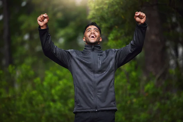 Young hispanic fit male athlete cheering with his fists in the air while on a run in a forest outside in nature. Exercise is good for health and wellbeing. Happy to reach his fitness goals.
