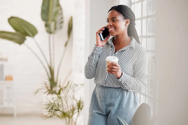 Young african american businesswoman on a call using her phone while drinking a coffee alone in an office at work. One black woman talking on her cellphone and holding a coffee cup on a break standing