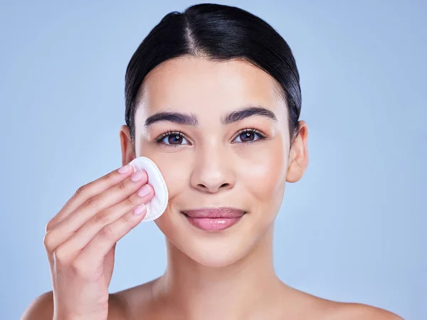 Studio Portrait of a beautiful mixed race woman using a cotton pad to remove makeup during a selfcare grooming routine. Hispanic woman applying cleanser to her face against blue copyspace background.
