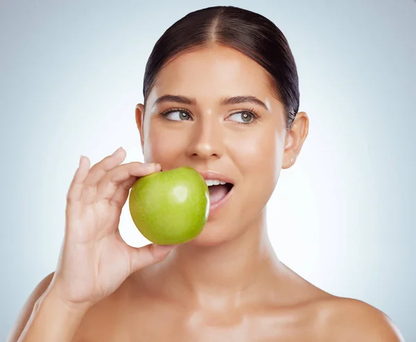 Beautiful woman biting into a green apple while posing with copypsace. Caucasian model looking contemplative while isolated against a grey studio background. Eating healthy is part of skincare routine